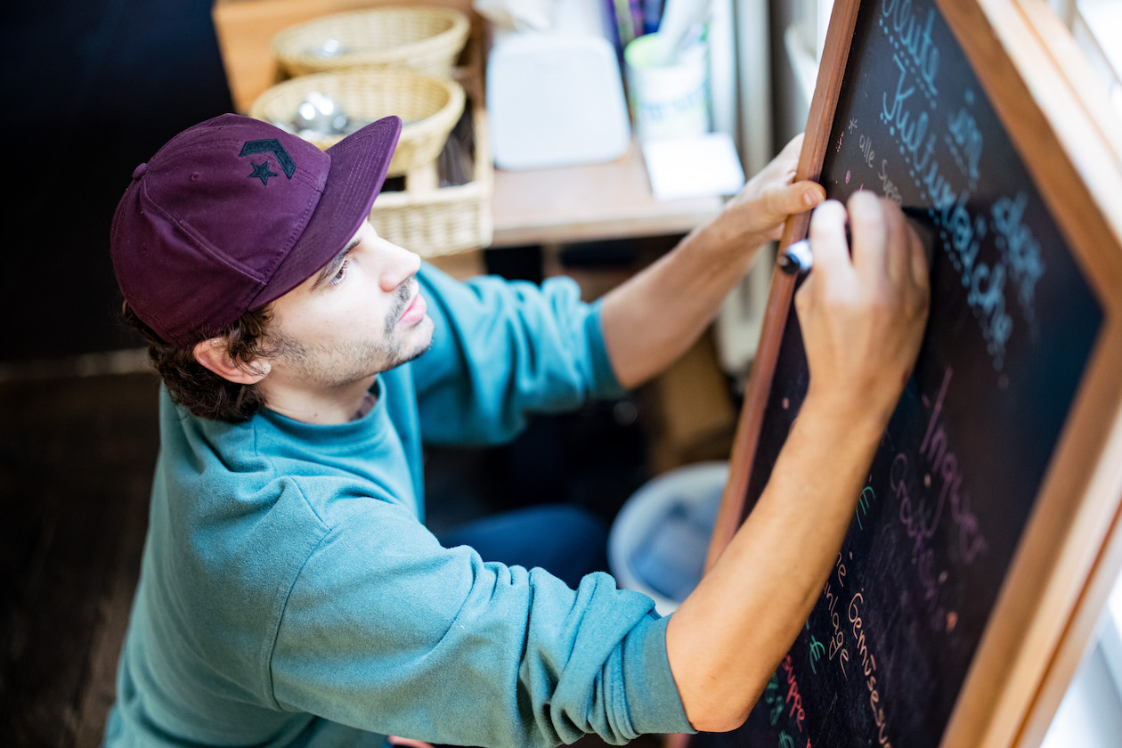 Ein junger mann in Türkise Pullover und mit einer violetten Cap beschriftet eine schwarze Tafel mit dem Mittagstisch.
