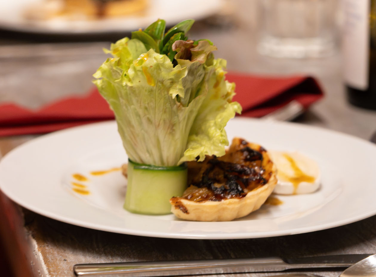 Teller mit einer kulinarischen Vorspeise. Man sieht eine gerollte Gurke mit Salatblatt und einen Zwiebelkuchen. Im Hintergrund sind Gläser zu sehen und eine rote Servierte.