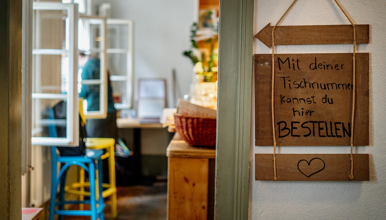 Auf einem Holzschild steht "Mit deiner Tischnummer kannst du hier bestellen". Daneben ist ein Türrahmen, im Hintergrund sind offene Fenster und zwei bunte Stühle zu sehen.