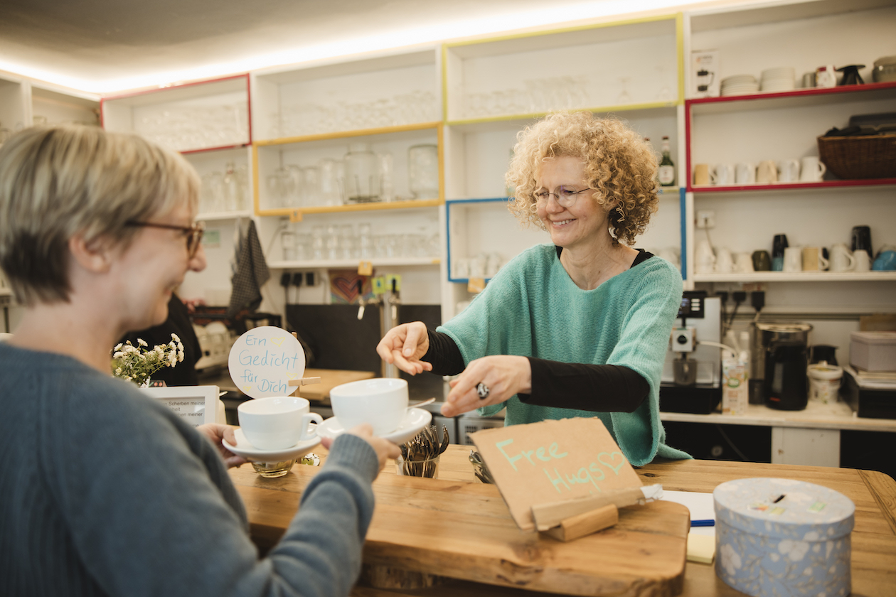 Zwei Frauen mit blonden Haaren, die an der Theke in der Kulturküche stehen und Kaffeetassen in der Hand haben. Im Hintergrund sieht man ein großes Regal mit Gläsern, Karaffen und mehr. Auf der Theke steht ein kleines Plakat mit der Aufschrift "Free Hugs".