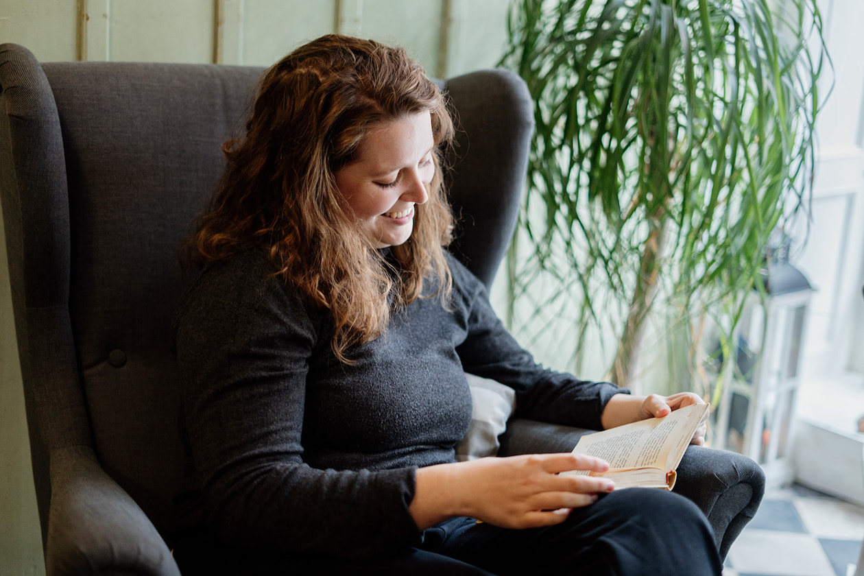Eine Frau mit Locken sitzt in einem grauen Sessel und liest. Im Hintergrund steht eine grüne Pflanze.