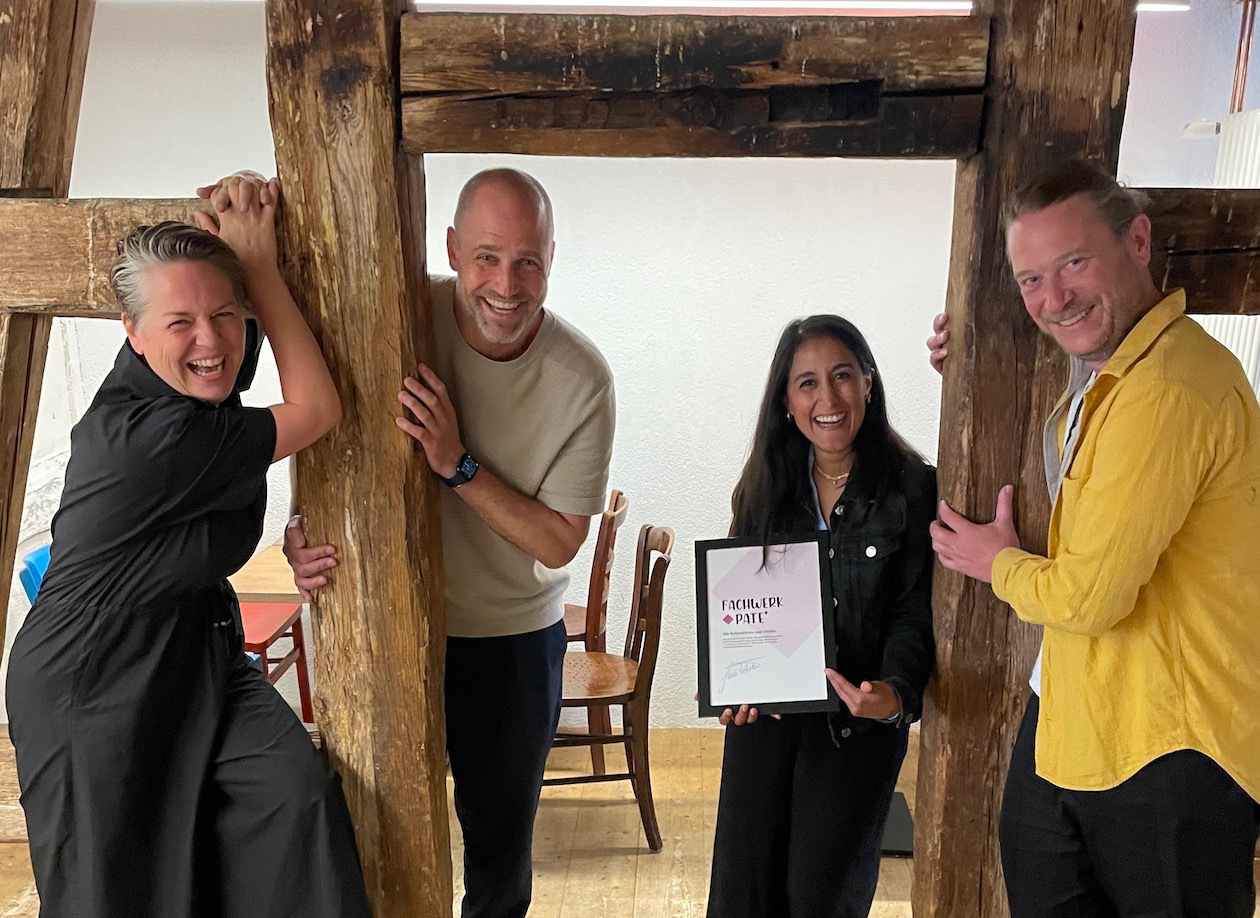 Vier Menschen, die lachen und sich am Fachwerkbauten festhalten. Die Frau in der Mitte hält eine Urkunde mit der Aufschrift "Fachwerkpate"