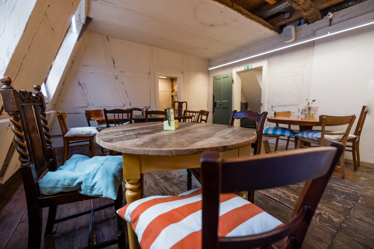 Im Obergeschoss der Kulturküche findet sich ein alter schiefer Holzboden. Außerdem stehen dort alte Holztische und Stühle. Im Hintergrund sieht man eine grüne Tür. Auf den Stühlen liegen bunte Kissen.