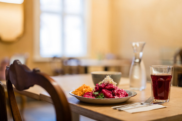 Tisch auf dem ein Teller mit Pasta steht, daneben ein Glas mit Traubenschorle, einer Wasser-Karaffe. Vor dem Tisch steht ein alter hölzerner Stuhl.
